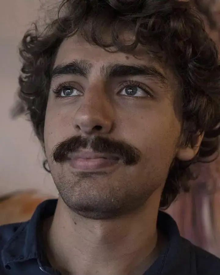 Portrait of Kaan Tulunay, a curly-haired man with a moustache, looking up and to the side